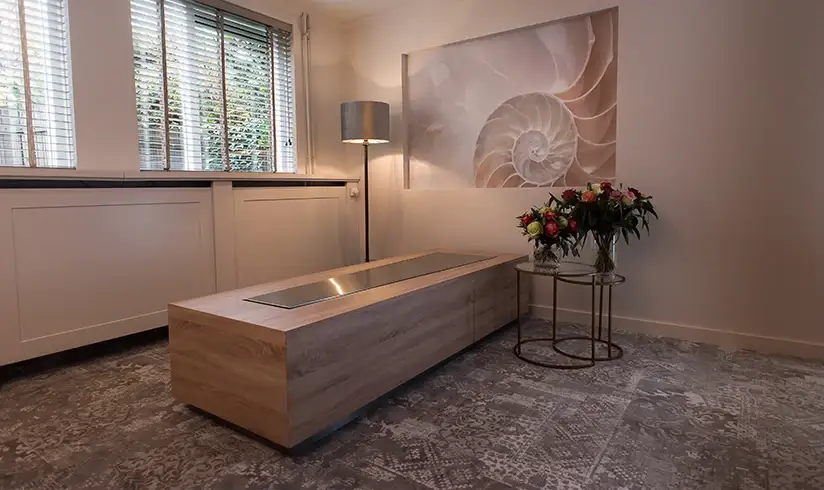 Een lichte kamer met een tafel voor een uitvaartkist en bloemen op een tafeltje